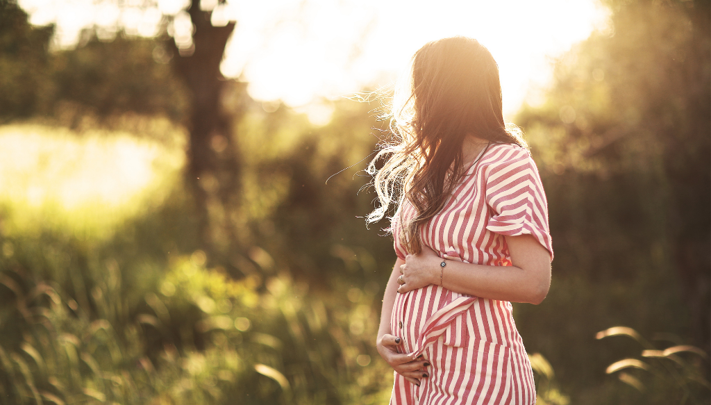 A pregnant traveller standing on a mountain trail, looking out over the landscape
